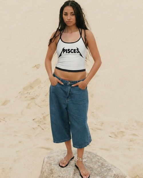 Eve's Essentials Pisces Equinox Tank campaign still — model standing on rock in warm sand landscape, cropped white tank and denim bermuda shorts